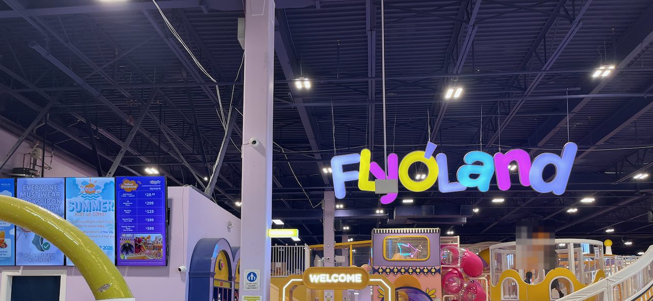 트와센 밀스 플라이오랜드 입구. 천장에 무지개색 FLYOLAND 네온 사인이 걸려 있고 왼쪽 벽면 스크린에 입장료와 패스 가격이 표시되어 있다
